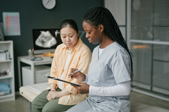 Asian pregnant young adult woman sitting on examination table listening to Black female medical professional explaining prenatal care while holding clipboard in modern clinic setting