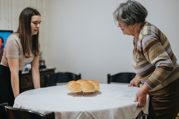 A senior woman and a younger female arrange a table with bread rolls, portraying inter generational...