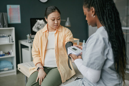 Pregnant Asian woman sitting on examination table receiving blood pressure check from Black female healthcare professional during prenatal care visit in medical office setting