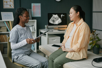 Asian pregnant woman sitting on examination table talking with Black female doctor during prenatal...