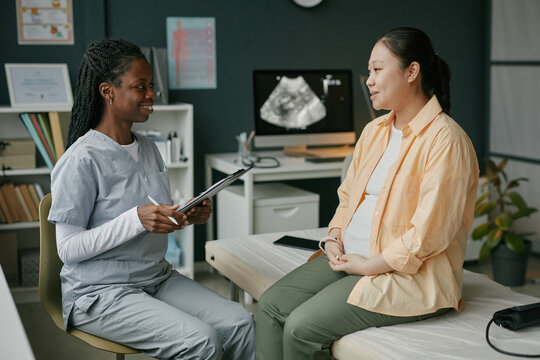 Asian pregnant woman sitting on examination table talking with Black female doctor during prenatal care appointment in medical office with ultrasound monitor visible in background - Powered by Adobe