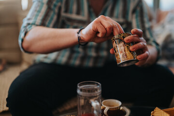 A close-up view of hands opening a beautifully decorated tin can in a comfortable home setting. The scene includes coffee cups and evokes feelings of warmth, comfort, and relaxation.
