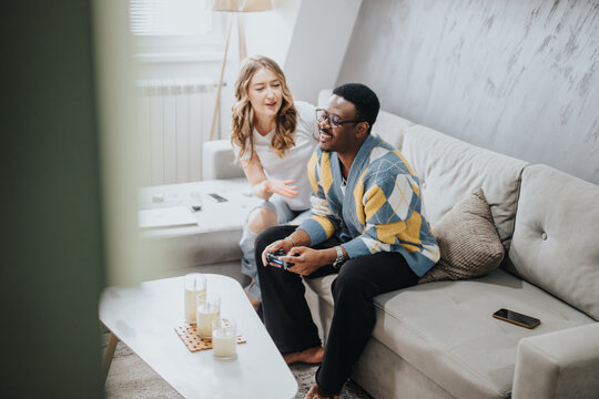 A happy couple spending their leisure time together at home, where the man plays a video game while the woman chats and laughs with him, creating a comfortable atmosphere.