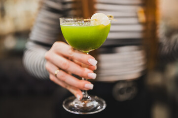 Close-up of a woman's hand holding a cocktail glass with green drink and lychee garnish, blurred background, elegant manicure, stylish and refreshing beverage presentation.