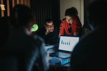 A group of business coworkers working together in a dimly lit office, focused on a laptop screen...
