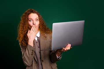 Naklejka premium Young surprised businesswoman with red curly hair using a laptop, wearing elegant suit, standing against green background