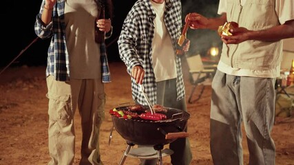 A group of Asian friends are enjoying a casual BBQ at night at a campsite. They are grilling skewers and drinking beer in front of their tents with a lively and relaxed atmosphere. 