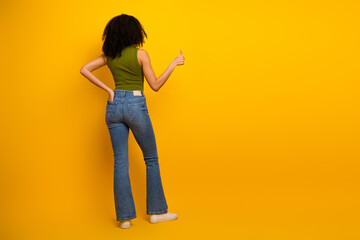 Female model in casual jeans and green top posing with thumbs up on vibrant yellow background