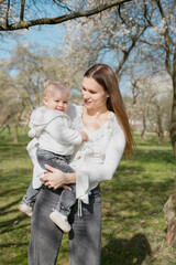 A beautiful mother and daughter look into the frame against the background of a blooming apple tree. A mother holds her little daughter in her arms. A spring day in the park.