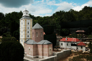 Obraz premium A detailed miniature of a church and traditional house displayed in an open-air miniature museum in Serbia. The scene features lush greenery and dramatic clouds above.