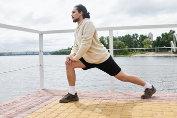 Young sporty man stretches outdoors by the water during a cloudy day