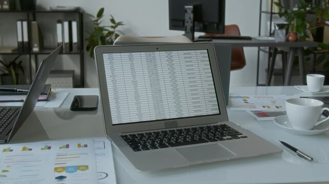 No people shot of white office desk featuring open laptop displaying detailed spreadsheet, surrounded by documents with charts, other devices, pen, coffee cups on saucers