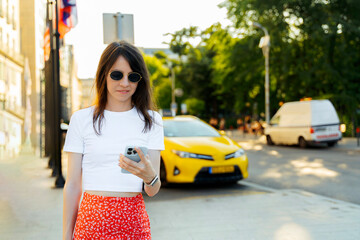 Young Woman Using Smartphone While Walking on City Street Near Yellow Taxi
