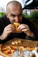 A man with a glass of dark beer in his hand enjoys a slice of pizza. There is a salt and pepper shaker and a kettle on the table.