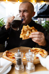 A man with a glass of dark beer in his hand enjoys a slice of pizza. There is a salt and pepper shaker and a kettle on the table.
