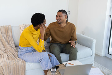 Laughing diverse couple sitting on light grey sofa in living room, with laptop and smartphone