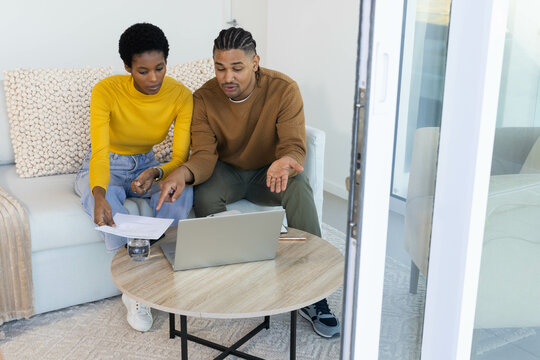 Reviewing diverse couple sitting on sofa in bright living room, with laptop documents pencil - Powered by Adobe