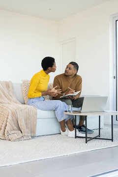Talking Diverse couple sharing notes on sofa in minimalist living room, with notebook and laptop