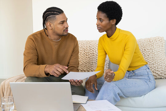 Reviewing documents Diverse couple sitting on sofa in living room, with laptop and notebook