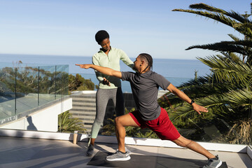Diverse trainer and client practicing yoga on seaside terrace, wearing sportswear with mat and cup