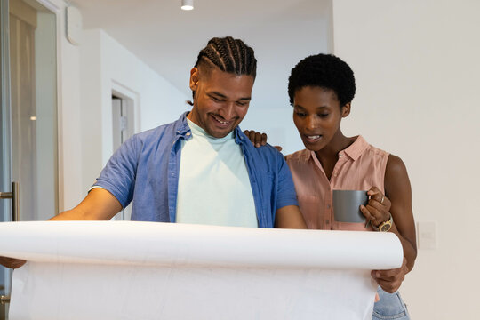 Reviewing diverse couple standing in modern hallway looking at large blueprint, with ceramic mug - Powered by Adobe