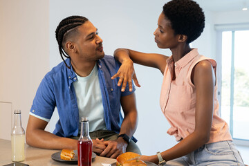Diverse couple eating burgers at quartz island counter in modern kitchen, with glass drink bottles