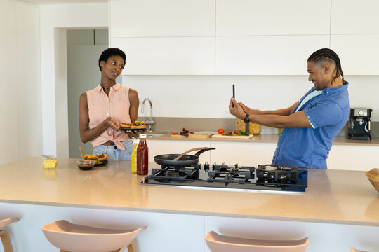 Presenting Diverse couple showing sandwiches on home kitchen island, with smartphone and toppings