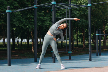young athletic caucasian woman doing stretching exercises outdoors in summer, grey sportswear,...