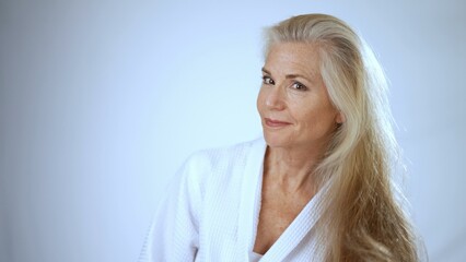A mature woman, dressed in a spa robe, smiles brightly as she brushes her long hair. The serene...