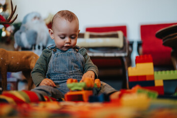 A toddler in overalls plays with bright construction toys in a cozy, holiday-decorated room. A...