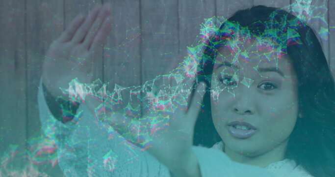 Interacting Asian woman in white sweater raising hands before wooden wall, with holographic network