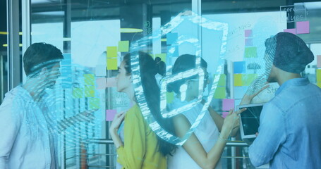 Collaborating team analyzing sticky notes on glass wall in office, with tablet and shield icon