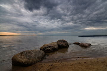 summer evening lake landscape. large coastal stones in calm water near sandy shore under cloudy dramatic sky with glow