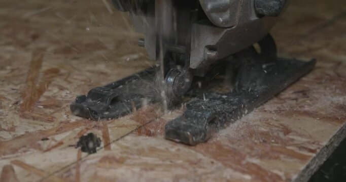 An experienced carpenter accurately cuts an OSB board with a jigsaw, chips fly around dynamically during woodworking in the workshop. Close-up of an electric jigsaw working on an OSB board.