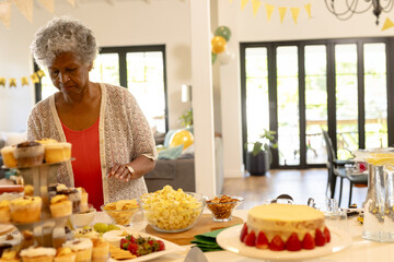 Senior African American woman selecting cupcakes cake popcorn at buffet table at home, copy space