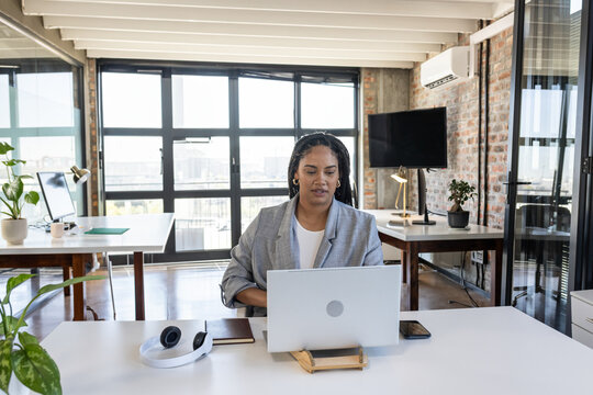 African American woman wearing blazer using laptop on stand at desk in open office with headphones