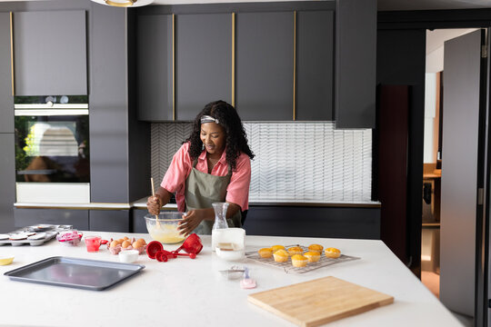 African American woman in green apron mixing batter in glass bowl at kitchen island with muffins