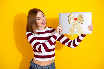 Smiling young woman in striped sweater holding a gift-wrapped laptop against a vibrant yellow...