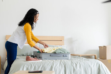 Person packing clothes into open suitcase on wooden bed with pink sneakers in tidy bedroom