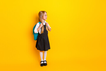 Charming elementary schoolgirl in uniform with backpack stands against bright yellow background,...