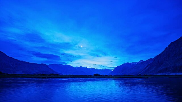 timelapse of Himalayas and blue sky mirrored in the Shyok River at sunrise, near lush Sumur village&mdash;where green orchards and barley fields thrive against Nubra Valleys stark mountain beauty.