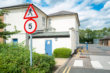Shallow focus of a speed limit and pedestrian crossing sign seen near a road crossing at a typical NHS hospital campus. Clinics are at the right of the image.
