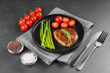 Tasty grilled beef steak served with green asparagus and cherry tomatoes on black plate, with cutlery. Perfect for food menu, diet, or culinary design concepts.