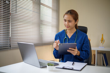 Female doctor in blue scrubs reviewing data on a tablet while sitting at a medical office desk.