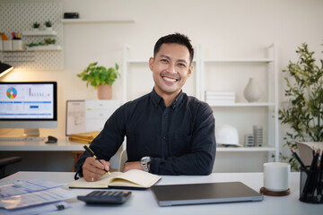 onfident young Asian businessman smiling at the camera while working at his desk in a modern office.