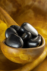Wooden bowl filled with black stones sits on a wooden table. The bowl is large and deep, and the stones are arranged in a neat pile. The stones are of various sizes and shapes