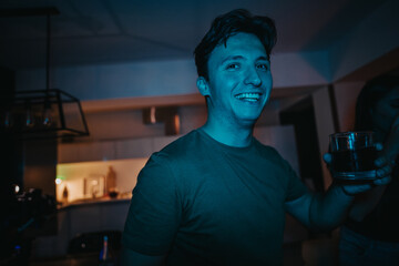 A joyful man holding a drink, enjoying an evening gathering with friends in a warmly lit room,...
