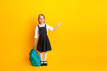 Smiling schoolgirl pointing with her finger while holding a backpack against a yellow background,...