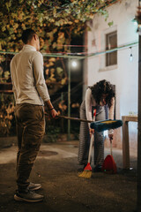 A man and a woman cleaning outside under soft evening lighting with brooms, surrounded by greenery...