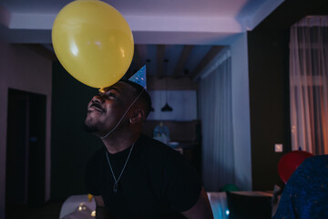 A joyful man wearing a party hat plays with a yellow balloon indoors at night.